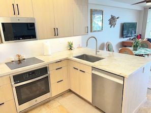 A modern kitchen with built-in appliances, a large island with a sink, and a dining area with a table and chairs. - Kona Alii 306 Oceanfront Corner Condo, Amazing View in the Heart of Kona (Kailua-Kona)