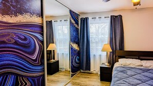 A bedroom with a large mirror, a bed with a blue quilt, two bedside tables with lamps, and a view through the window. - Nice and cozy duplex close to everything in KC! (Overland Park)