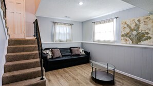 A living room with a staircase, a black sofa, a glass coffee table, and a wooden floor. - Nice and cozy duplex close to everything in KC! (Overland Park)
