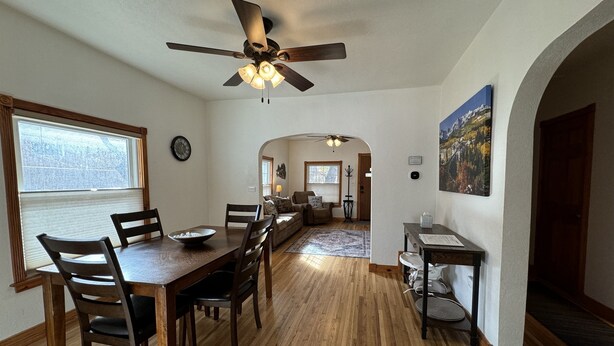 Un espace repas avec une table et des chaises en bois, un ventilateur de plafond et une vue sur la cuisine à travers une arche.