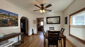 Un coin repas avec une table et des chaises en bois, un ventilateur de plafond, un grand miroir et une fenêtre avec des stores.