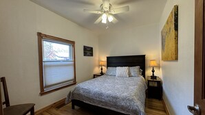Une chambre à coucher comprenant un lit, deux tables de chevet, un ventilateur de plafond, une fenêtre avec des stores et une porte.