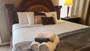 A neatly made bed with a wooden headboard, white and brown pillows, a rolled towel, and a bedside table with a lamp.