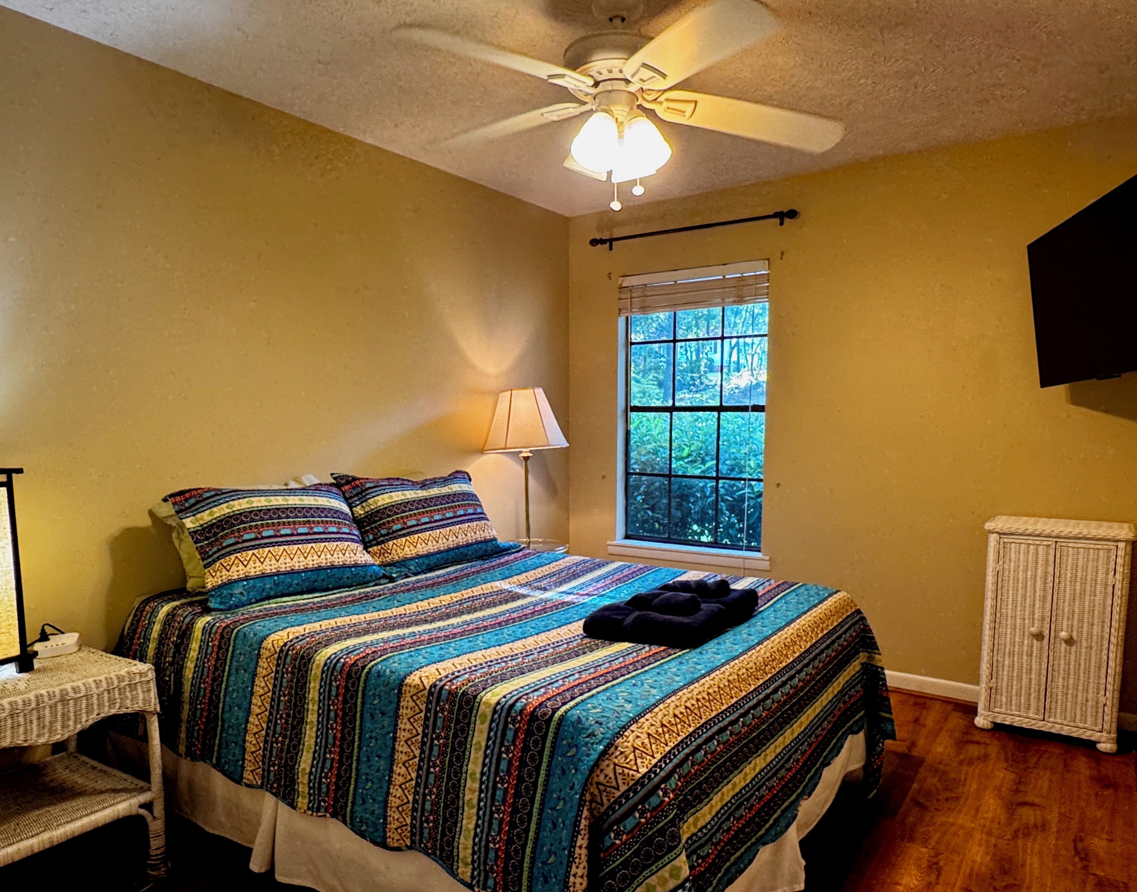 A bedroom with a bed, a window, a ceiling fan, a bedside table, and a cabinet.