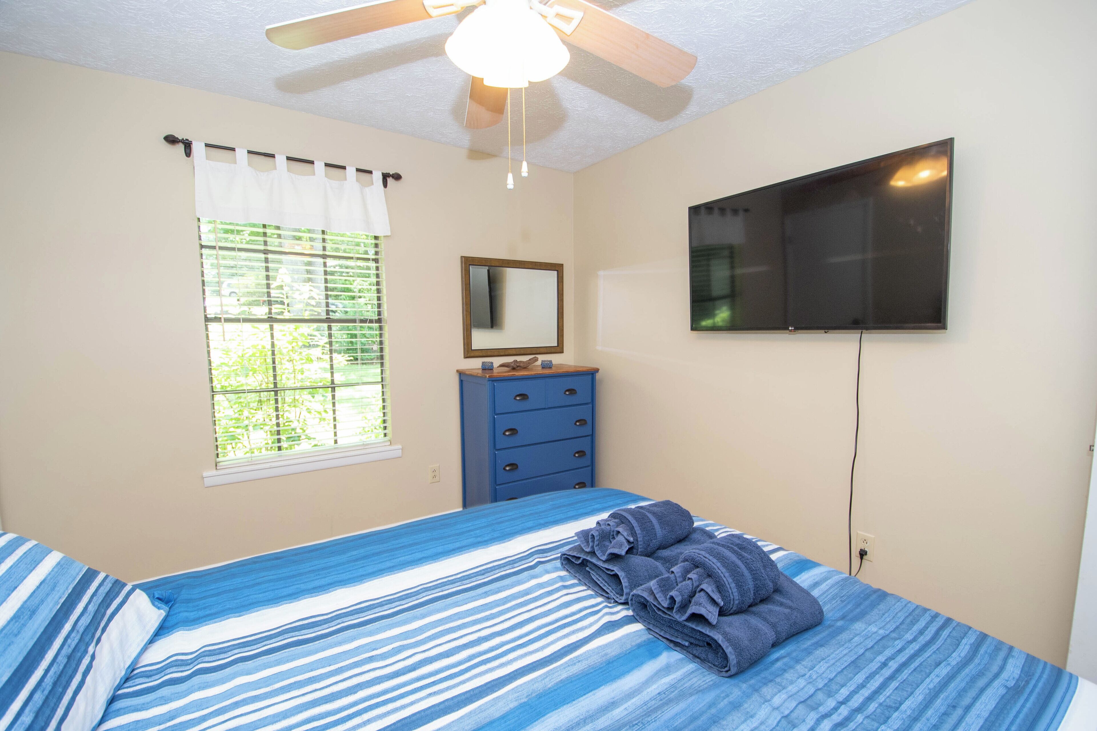 A bedroom with a bed, a dresser, a ceiling fan, a TV, and a window with blinds.