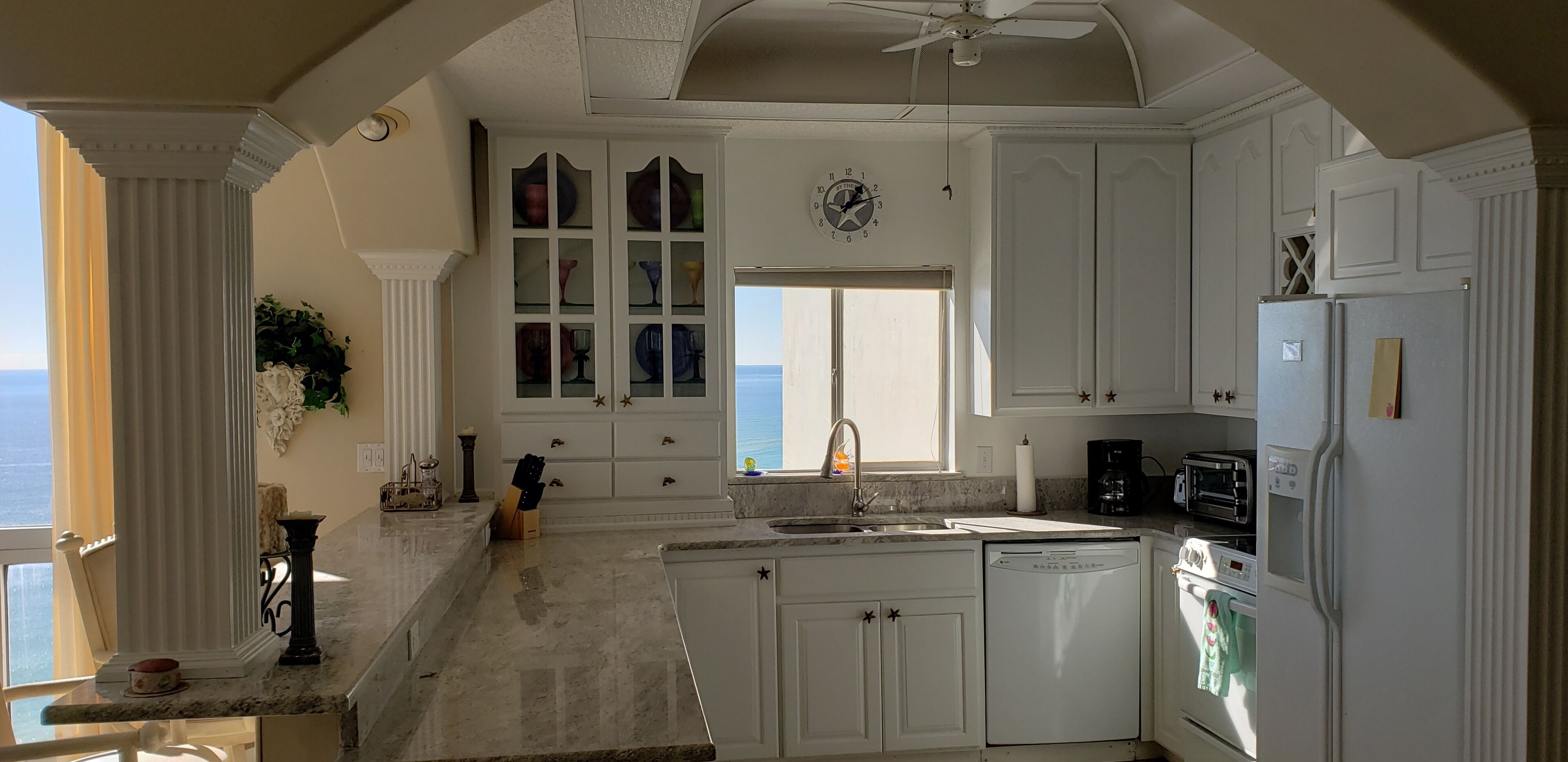 Uma cozinha com armários brancos, forno embutido, lava-louças e uma janela com vista para o oceano.