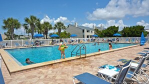 A swimming pool with lounge chairs and umbrellas, surrounded by palm trees and a building in the background.