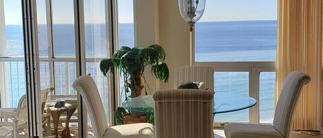 Ruime kamer met groot raam en uitzicht op de oceaan. Er is een rieten bank, een ronde tafel en twee stoelen.