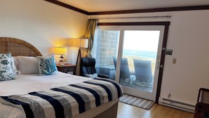 Habitación de hotel con una cama grande, una mesita de noche, una lámpara, una silla y vista al océano a través de una puerta corrediza de vidrio.