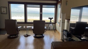 Una habitación con ventanas amplias que dan al océano, dos sillas, un telescopio y un escritorio con una computadora.