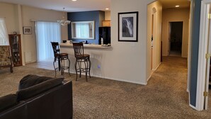 A living room with a dark brown sofa, a small bar area with two stools, a chandelier, and a large window with blinds. - Tri-Cities Home Away From Home (Richland)