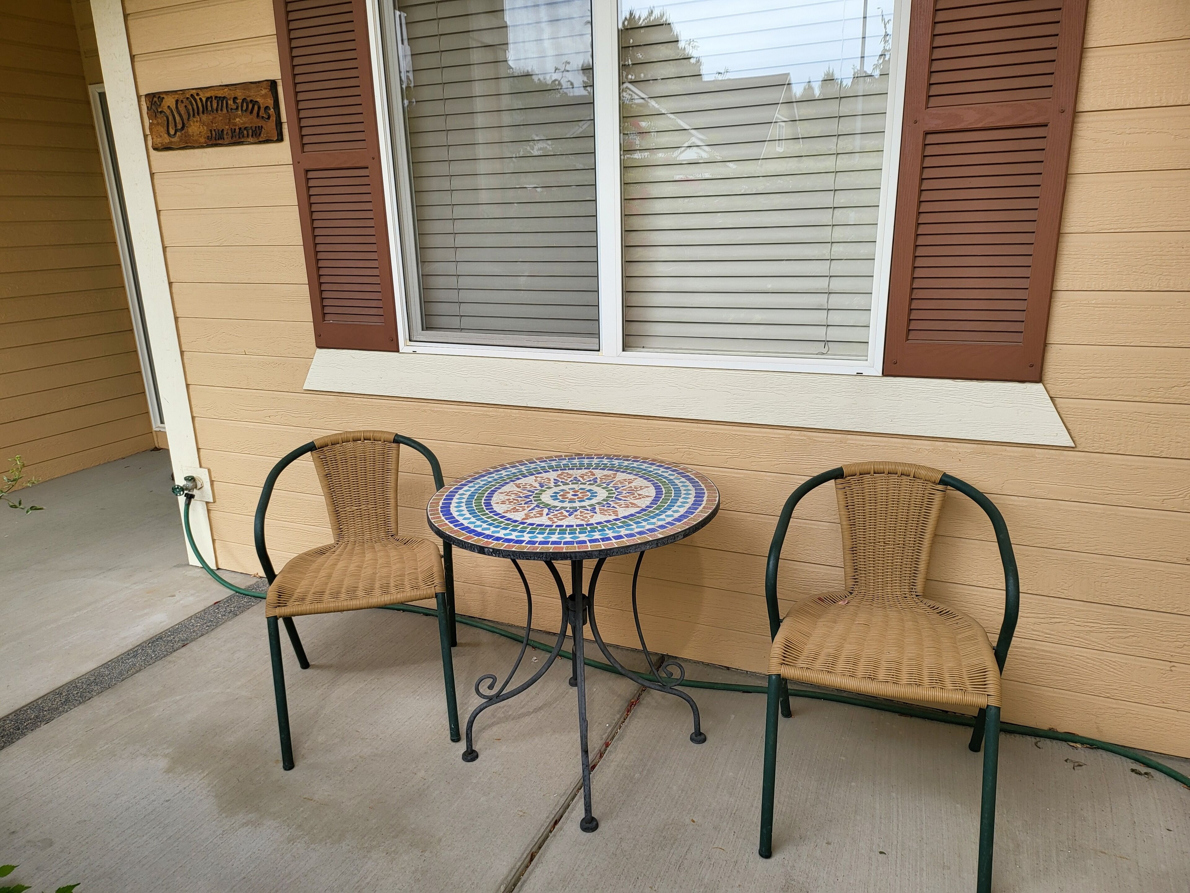 A small outdoor seating area with two wicker chairs and a mosaic-tiled table, positioned under a window with brown shutters.