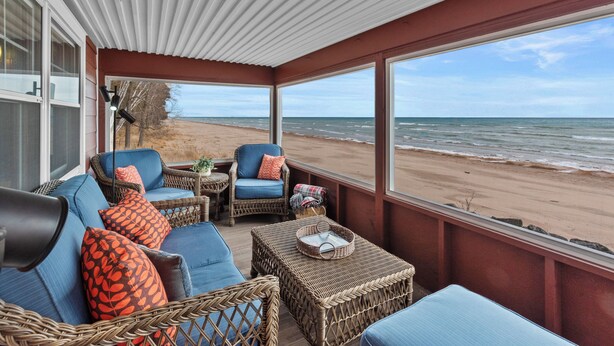 A covered outdoor seating area with wicker furniture, a view of the beach, and a large window. - Private Beach, Outdoor Dining, Firepit (Oostburg)