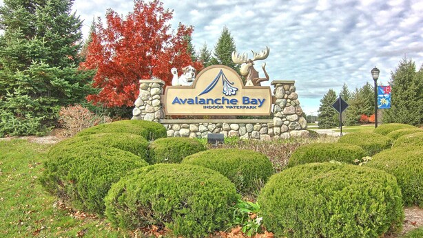 A sign for Avalanche Bay Indoor Waterpark with a moose and dog logo, surrounded by well-manicured bushes and trees.