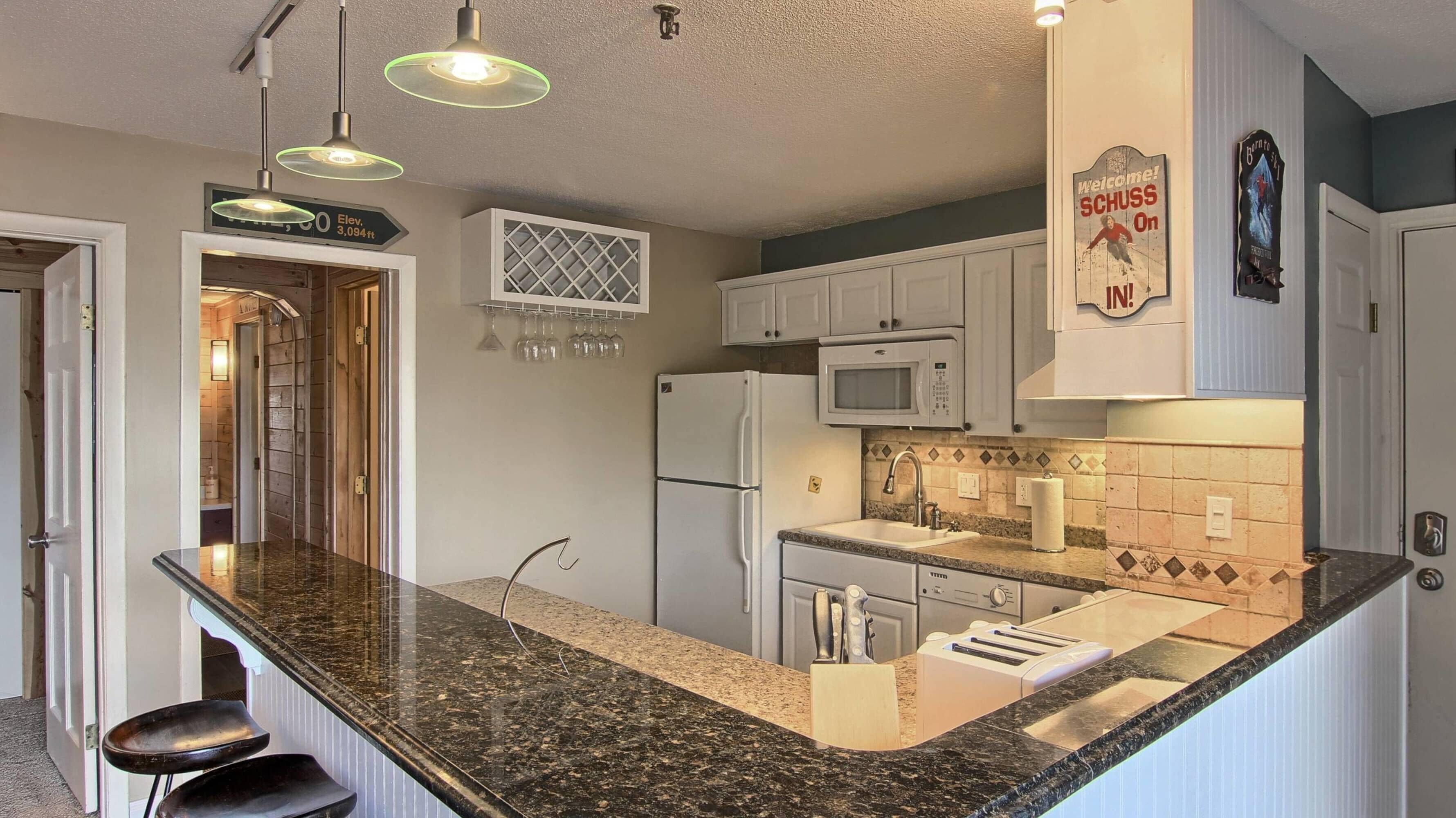 Una cocina con encimeras de granito, armarios blancos, un refrigerador, un microondas y un tostador. Hay luces colgantes y un letrero sobre el lavamanos.