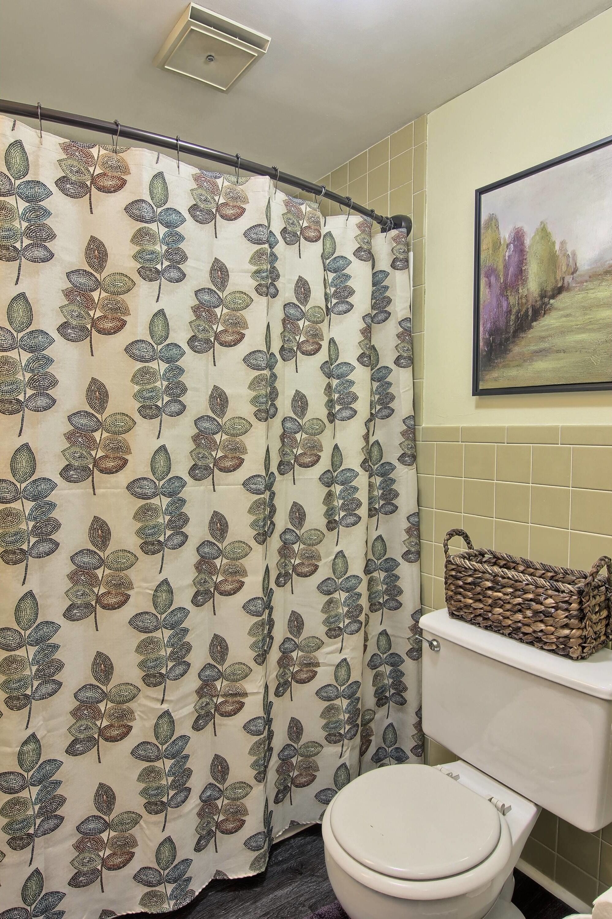 Ein Badezimmer mit Duschvorhang mit Blättermotiv, Toilette, einem gewebten Korb an der Wand und einem gerahmten Bild über der Toilette.