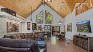 A spacious living area with a high ceiling, a large window, a fireplace, a dining area, and a television. - Large, Beautiful Cabin on Tonto Creek in Kohl's Ranch /Game Room/Hot Tub/Views (Payson)