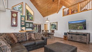A spacious living room with a leather sofa, ottoman, wooden ceiling, large windows, dining area, and a TV. - Large, Beautiful Cabin on Tonto Creek in Kohl's Ranch /Game Room/Hot Tub/Views (Payson)