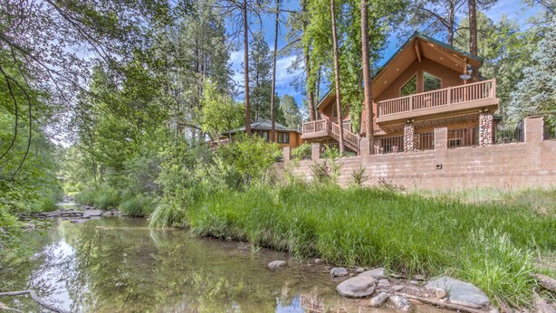 A lodge with a stone exterior, a balcony, and a view of a forested area with a stream. - Large, Beautiful Cabin on Tonto Creek in Kohl's Ranch /Game Room/Hot Tub/Views (Payson)