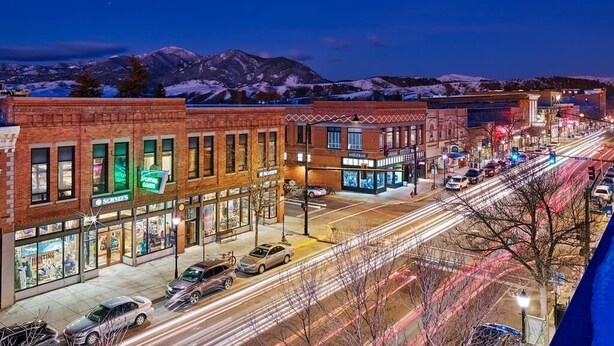 A street view at dusk with buildings, a mountain range in the background, and light trails from vehicles.