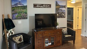 A living room with a flat-screen TV, wooden entertainment center, two leather armchairs, and wall-mounted artwork depicting Montana landscapes.