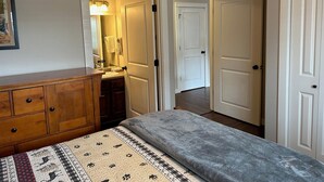 A bedroom with a wooden dresser, a bed with a patterned blanket, a bathroom with a mirror and towel rack, and white doors leading to other rooms.