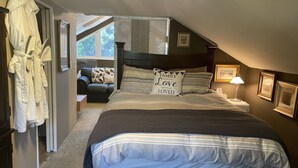 Une chambre à coucher avec un grand lit, une table de chevet avec une lampe, une fenêtre avec des stores, une chaise et un porte-manteau.