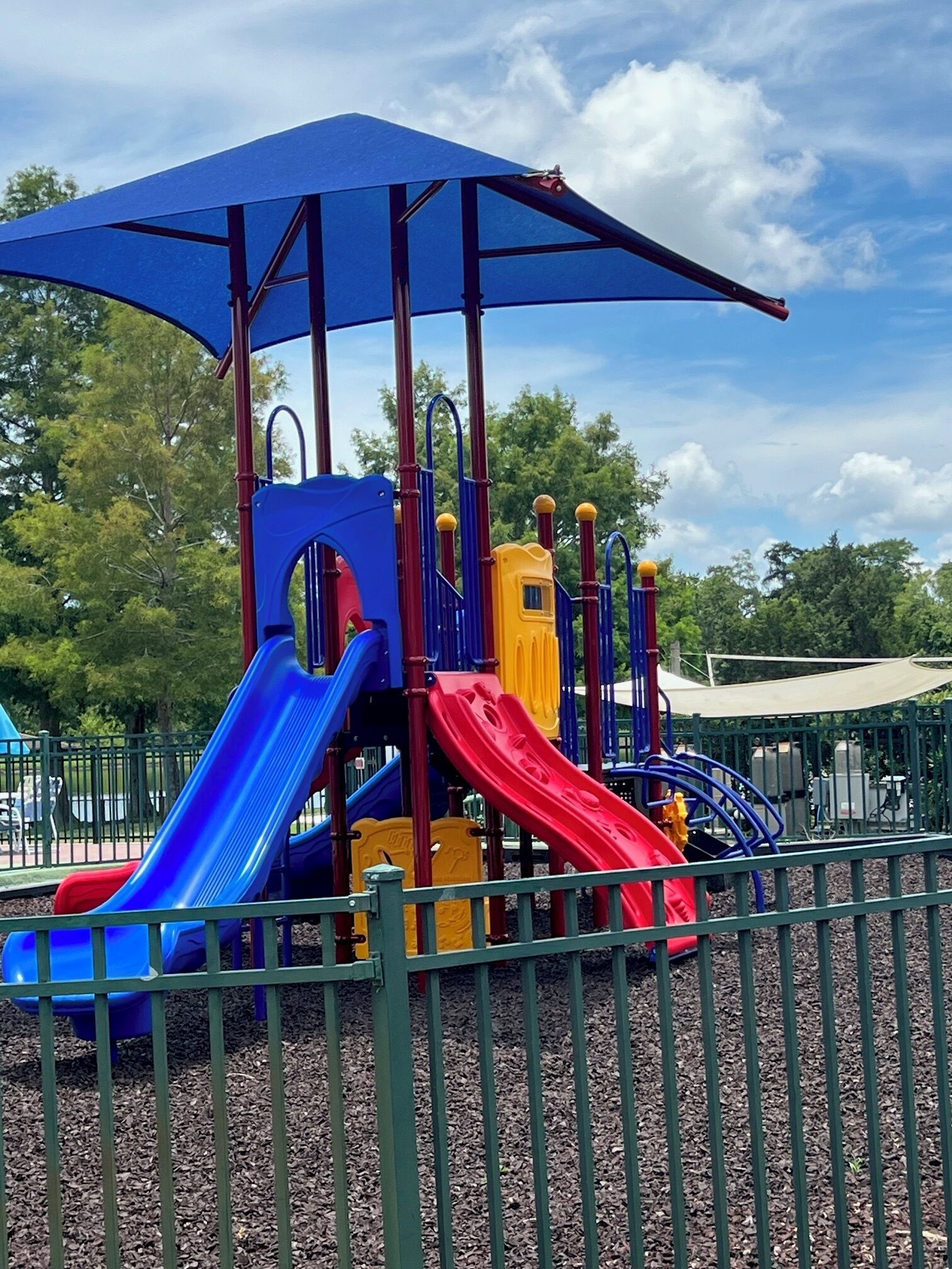 Un parque infantil con toboganes y estructuras para escalar, rodeado por una cerca y árboles.