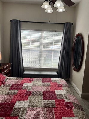 Een slaapkamer met een groot bed, een raam met zonwering, een plafondventilator en een spiegel aan de muur.