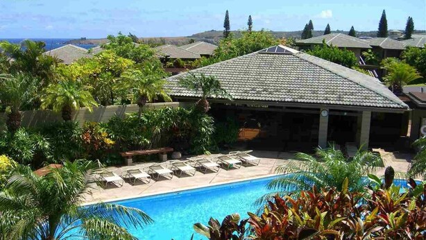 A pool area with lounge chairs, surrounded by tropical plants and a building with a tiled roof. - Condo in Kapalua!! (Maui)
