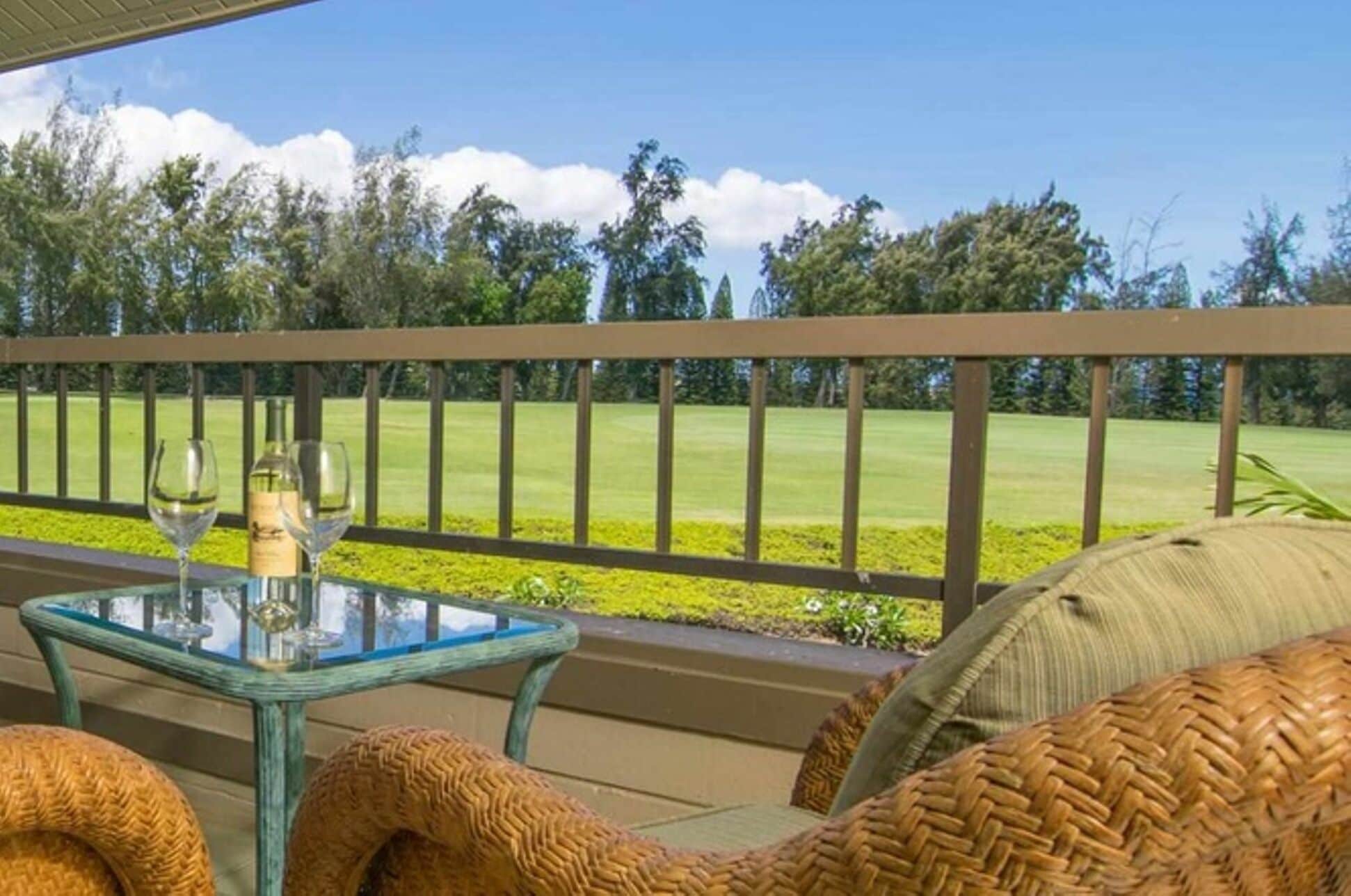 A glass-top table with two wine glasses and a bottle, a wicker chair, and a view of a golf course.