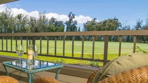 A glass-top table with two wine glasses and a bottle, a wicker chair, and a view of a golf course. - Condo in Kapalua!! (Maui)