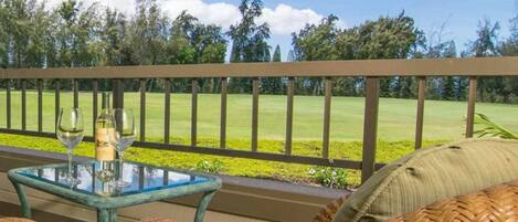 A glass-top table with two wine glasses and a bottle, a wicker chair, and a view of a golf course.