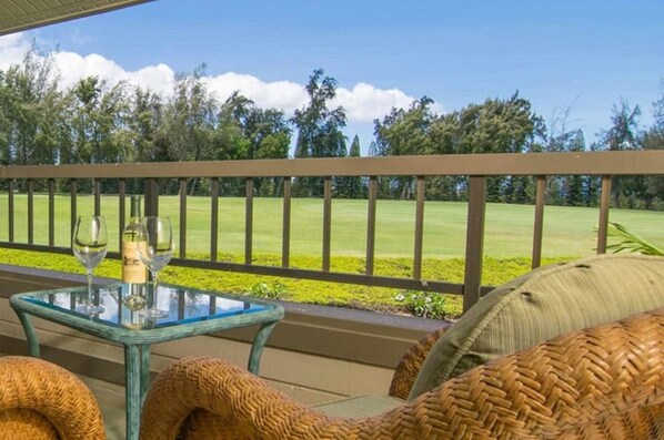 A glass-top table with two wine glasses and a bottle, a wicker chair, and a view of a golf course.