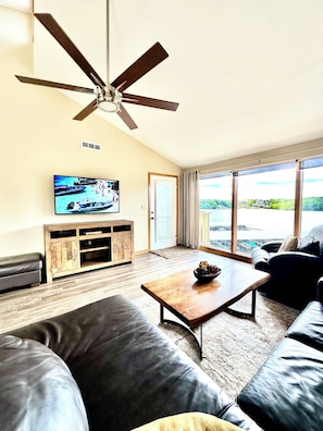 Relax in this spacious living room with a flat-screen TV and a comfortable leather sofa, offering a view of a serene body of water and lush greenery.