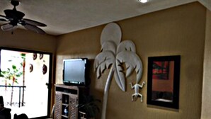 A living room with a television on a wooden stand, a ceiling fan, a decorative palm tree, and a small table with a chair.