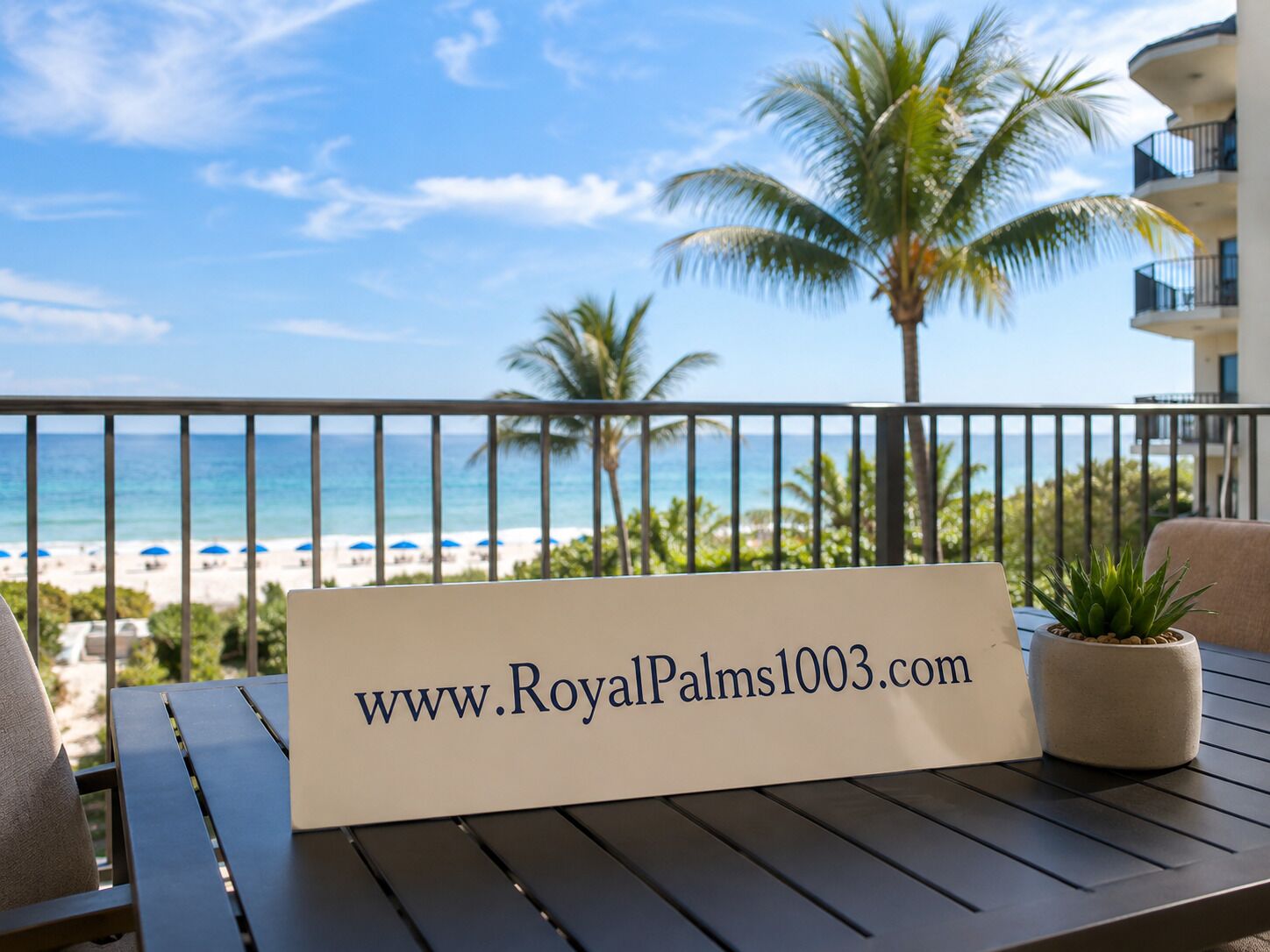 A balcony with a sign displaying a website address, overlooking a beach with palm trees and a clear sky.