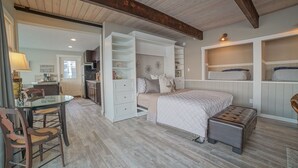 Quarto com cama, estantes embutidas, área de refeições com mesa e cadeiras, e cozinha ao fundo.