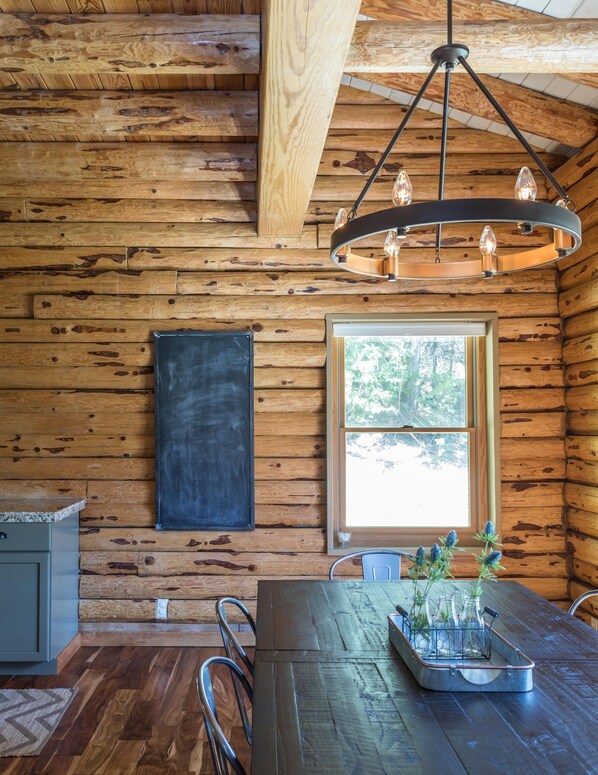 Interior rústico de una cabaña de madera con una mesa de comedor, sillas, una pizarra y una ventana con vistas a árboles.
