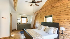 Une chambre avec un mur en rondins de bois, un grand lit avec une literie à motifs, un ventilateur de plafond et une petite table de chevet avec une lampe.