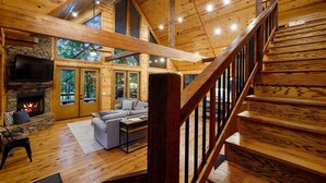 Un interior espacioso de madera con chimenea, una escalera y un amplio ventanal con vistas a la vegetación.
