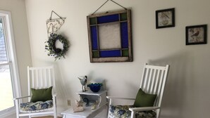 Two white rocking chairs with green cushions, a small white table with decorative items, and a wall with a blue-framed artwork and two framed pictures.