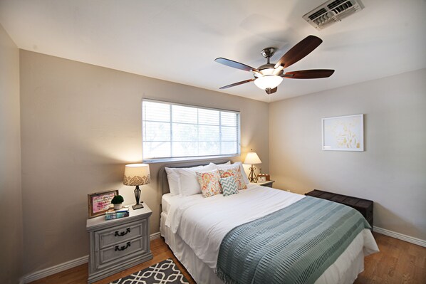 A bedroom with a bed, two bedside tables, a ceiling fan, a window, and a wall-mounted artwork.