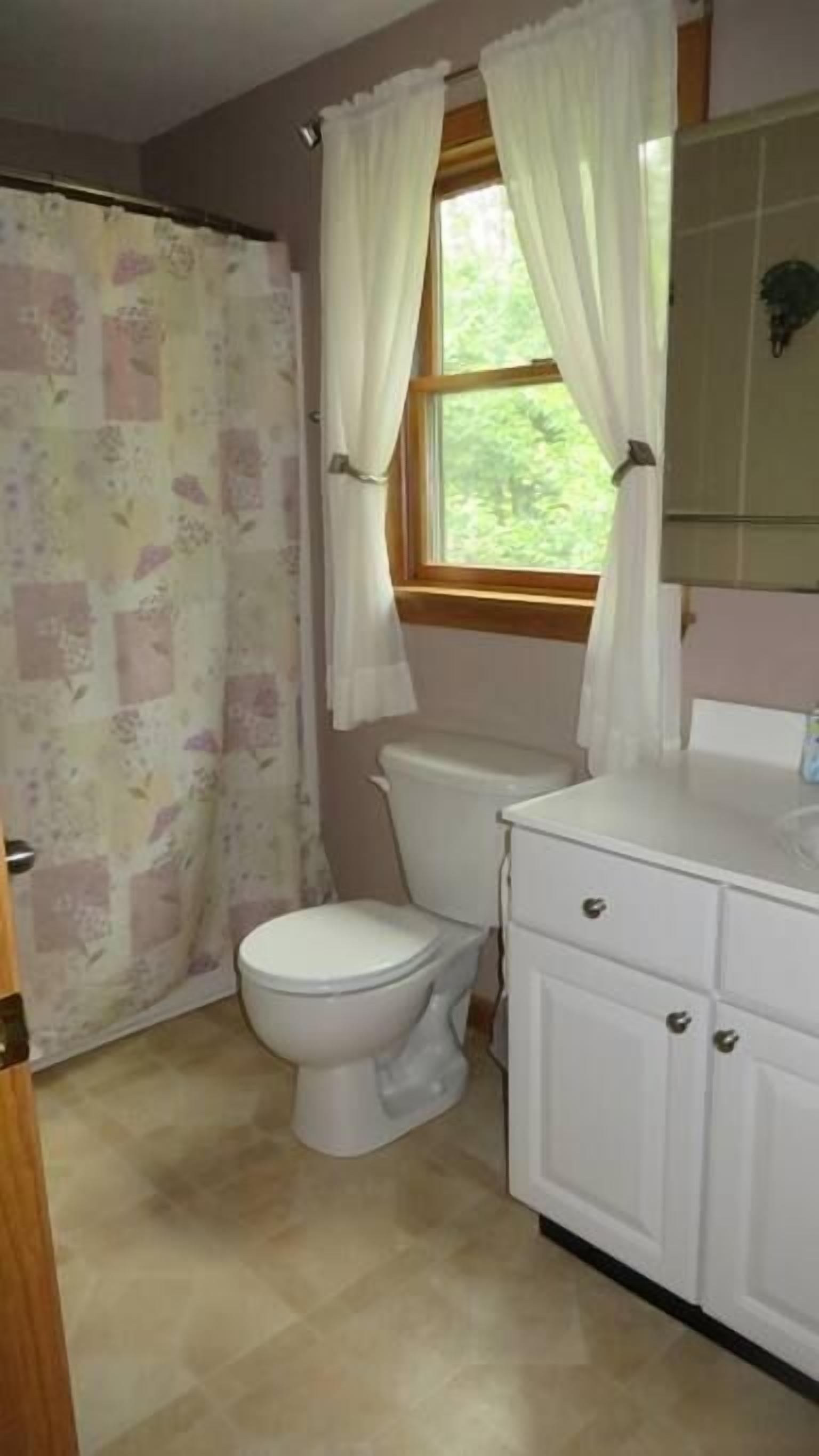 Een badkamer met toilet, douchegordijn en een raam met gordijnen.