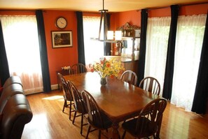 A dining room with a wooden table, six chairs, a vase of flowers, a clock, and a window with curtains. - Eagles Nest at Loon Lake near Gore Mountain (Chestertown)