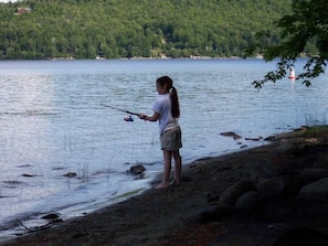 A person fishing by a lake with a forested hill in the background.