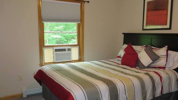 Un dormitorio con cama, una ventana con persianas, un aire acondicionado instalado en la pared y un cuadro enmarcado sobre la cama.