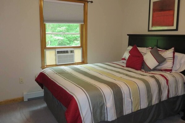 A bedroom with a bed, a window with blinds, a wall-mounted air conditioner, and a framed picture above the bed.