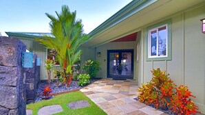 A house with a stone pathway leading to a dark-colored door, surrounded by a well-maintained garden with various plants and a palm tree.