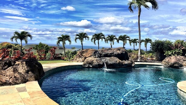 A pool with clear blue water, surrounded by rocks and palm trees, with a view of the ocean and sky.
